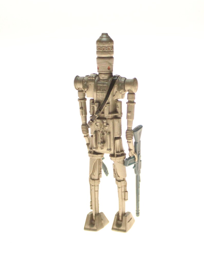 May include: A beige and silver action figure of a Star Wars character, a droid, holding a blue and gray blaster rifle. The droid has a tall, cylindrical head with a small, round face.
