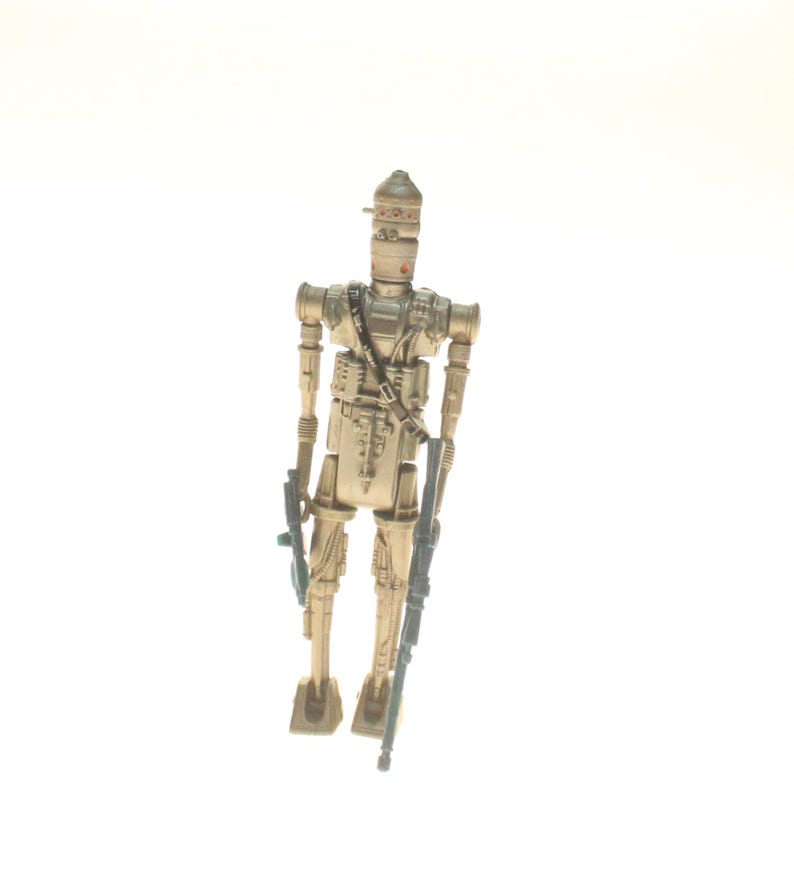 May include: A beige and brown plastic action figure of a robot with a helmet and two guns. The robot is standing on a white background.