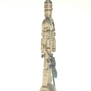 May include: A white and gray action figure of a Star Wars character, a droid with a large head and a weapon in its hand. The figure is standing on a white background.