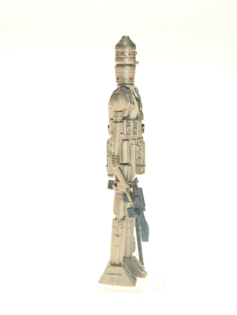 May include: A white and gray action figure of a Star Wars character, a droid with a large head and a weapon in its hand. The figure is standing on a white background.