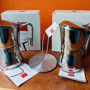 Op de afbeelding: Twee glanzende espressomakers van roestvrij staal, een standaard en dozen. De espressomakers hebben een klassiek ontwerp met een handvat en een tuit. De dozen zijn wit met rode accenten. De standaard is zilverkleurig en cirkelvormig.