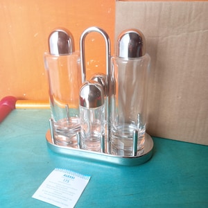 Pode incluir: Um conjunto de condimentos em aço inoxidável com três recipientes de vidro transparente e tampas prateadas. O conjunto inclui um saleiro, um pimenteiro e duas garrafas, todos presos em um suporte de metal prateado. Um pequeno cartão com o texto "ALESSI 175" está em primeiro plano.