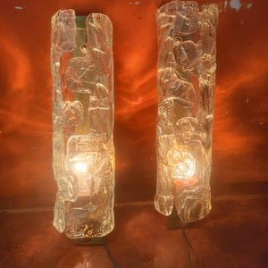 May include: A pair of wall sconces featuring vertical, cylindrical glass shades with a textured, organic, ice-like surface. Each sconce is mounted on a metallic backplate and contains a single illuminated light bulb, casting a warm glow.