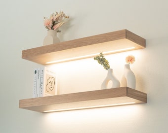 May include: Two wooden shelves with a white background. The top shelf has a small white vase with dried flowers. The bottom shelf has a stack of books and two white vases with flowers.