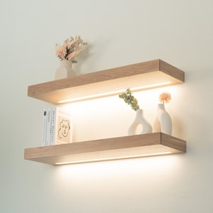 May include: Two wooden shelves with a white background. The top shelf has a small white vase with dried flowers. The bottom shelf has a stack of books and two white vases with flowers.