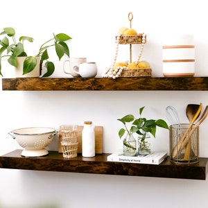 May include: Two dark stained wooden floating shelves against a white wall. The top shelf holds plants, mugs, and a tiered basket with fruit. The bottom shelf displays a colander, glasses, a bottle, and a book.
