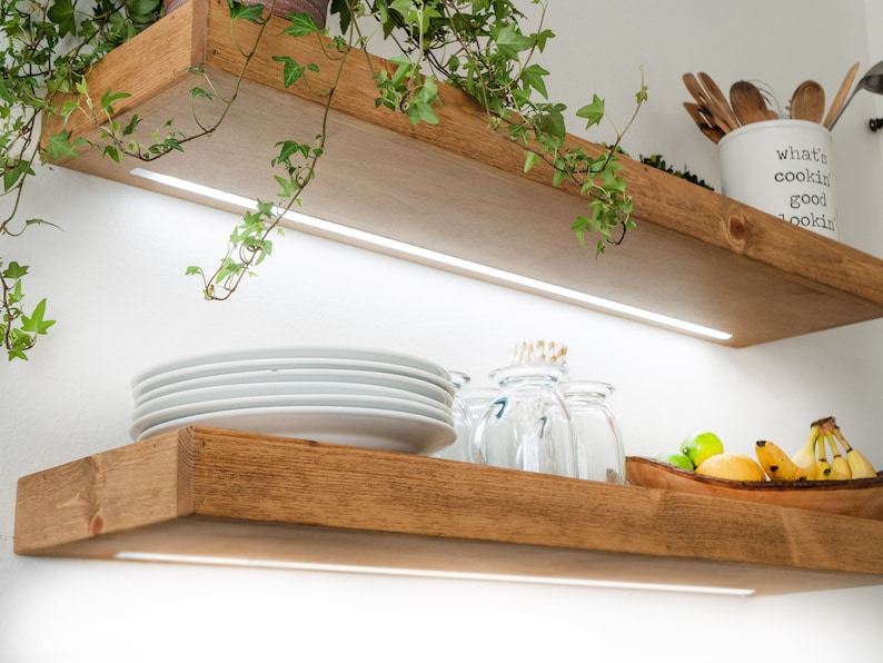 May include: Two wooden shelves with a white background. The top shelf has a potted plant with green leaves hanging over the edge. The bottom shelf has a stack of white plates, a glass jar, and a bowl of fruit. Both shelves have white LED lights underneath.