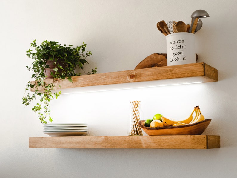 May include: Two wooden shelves with a white background. The top shelf has a plant in a brown pot, a wooden bowl, and a white ceramic utensil holder with the text "What's cookin' good lookin'". The bottom shelf has a stack of white plates, a glass vase with straws, a wooden bowl with bananas, limes, and lemons, and a white light strip.