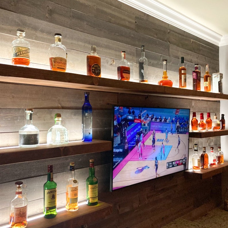 May include: A wooden shelf unit with multiple shelves, displaying a variety of liquor bottles. The shelves are lit with LED lights. A television screen is mounted on the wall above the shelves, showing a basketball game.