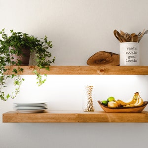 May include: Two wooden shelves with a plant, a white ceramic jar with the text "what's cookin' good lookin'" and utensils, a bowl of fruit, and a stack of white plates.