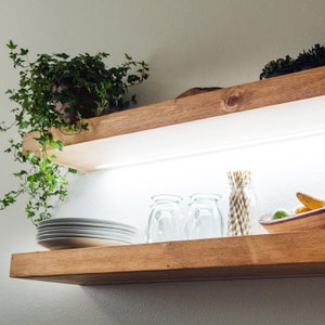 May include: Two wooden shelves with a light wood finish. The top shelf has a potted plant with green leaves. The bottom shelf has four clear glass cups, a bowl with fruit, and a bundle of straws.