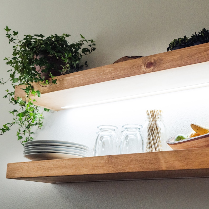 May include: Two wooden shelves with a light wood finish. The top shelf has a potted plant with green leaves. The bottom shelf has four clear glass cups, a bowl with fruit, and a bundle of straws.