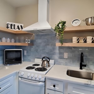 Op de afbeelding: Een witte keuken met lichtblauwe tegelachterwand, witte kasten en een wit fornuis. Boven het fornuis bevinden zich twee houten planken met een zilveren waterkoker, mokken en een klok. De planken zijn gelabeld met "RELAX".