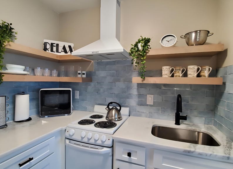 Op de afbeelding: Een witte keuken met lichtblauwe tegelachterwand, witte kasten en een wit fornuis. Boven het fornuis bevinden zich twee houten planken met een zilveren waterkoker, mokken en een klok. De planken zijn gelabeld met "RELAX".