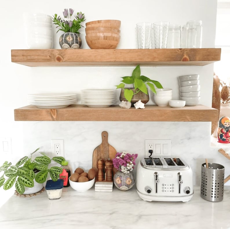 K&ouml;nnte beinhalten: Zwei Holzregale mit verschiedenen Gegenst&auml;nden darauf, darunter Pflanzen, Geschirr, Schalen und Gl&auml;ser. Die Regale sind an einer wei&szlig;en Wand &uuml;ber einer wei&szlig;en Arbeitsplatte mit einem Toaster, einem Schneidebrett und anderen K&uuml;chenutensilien angebracht.