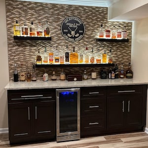 May include: A home bar with dark brown cabinets, a stainless steel mini-fridge, and two shelves with a variety of liquor bottles. The back wall is covered in a brown mosaic tile and features a round wooden sign with the text "The Jeffersons Melissa & Joe's 1978 Hole Est. 2014 Lake Ridge, Virginia".