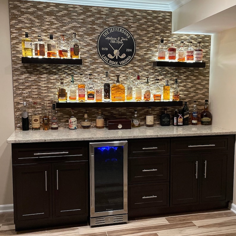 May include: A home bar with dark brown cabinets, a stainless steel mini-fridge, and two shelves with a variety of liquor bottles. The back wall is covered in a brown mosaic tile and features a round wooden sign with the text "The Jeffersons Melissa & Joe's 1978 Hole Est. 2014 Lake Ridge, Virginia".