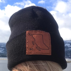 May include: Black knit beanie with a brown leather patch featuring the outline of British Columbia, Canada, and the letters "BC." The hat is worn outdoors with a scenic background of mountains and water.
