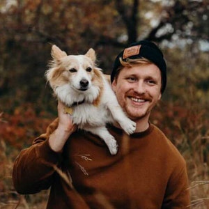May include: A smiling person wearing a brown sweater and a black beanie with a patch, holding a small, fluffy dog with tan and white fur. The dog is perched on the person's shoulder. The background is blurred with autumn colors.