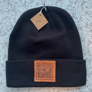 May include: Black beanie with a folded brim and a brown leather patch. The patch features an etched design of a helicopter, mountains, and the text "Nee&mmm...". A small tag with the word "HOME" hangs from the beanie.