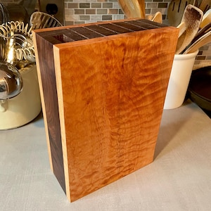 Könnte beinhalten: Ein Messerblock aus Holz mit einer satten, rotbraunen Oberfläche. Der Block hat eine kontrastierende dunkelbraune Seitenwand und Schlitze zur Aufbewahrung von Messern. Die Holzmaserung ist sichtbar und trägt zur natürlichen Ästhetik bei. Der Block ist rechteckig und steht aufrecht.