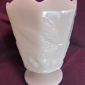 May include: A white milk glass vase with a scalloped rim and a footed base. The vase has a raised floral design.