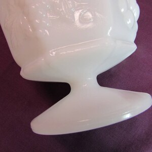 May include: White milk glass vase with a pedestal base and a floral design. The vase is made of opaque white glass and has a smooth, glossy finish.