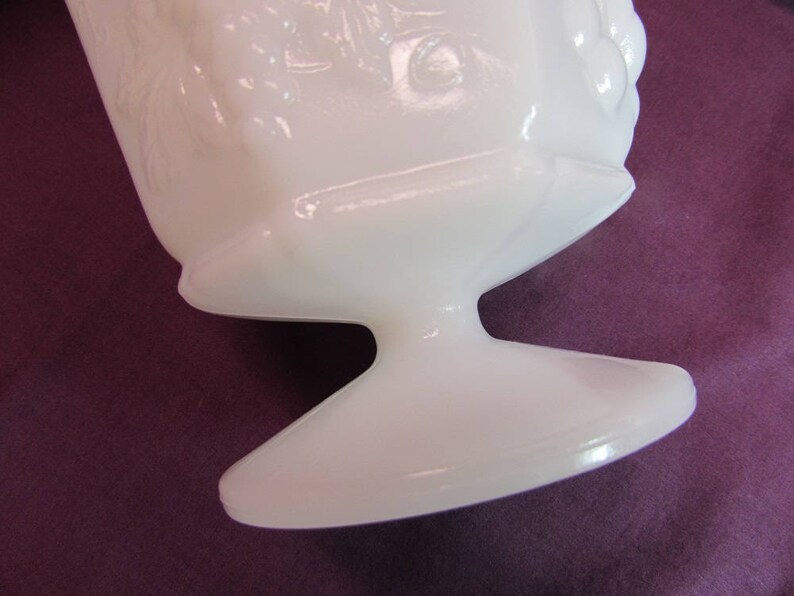 May include: White milk glass vase with a pedestal base and a floral design. The vase is made of opaque white glass and has a smooth, glossy finish.