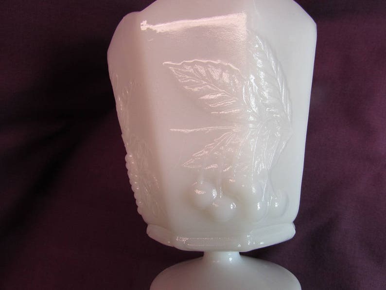 May include: White milk glass pedestal bowl with an embossed design of leaves and berries. The bowl has a scalloped rim and a pedestal base.