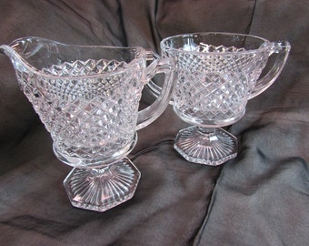 May include: A set of two clear glass creamer and sugar bowls with a diamond pattern and a pedestal base.