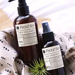 May include: Two brown Pantry brand pet care products: a Dirty Dog Wash bottle with a pump and a Dog Deodorant spray bottle. Both have white labels with black text. The bottles are on a patterned cloth with a small plant.