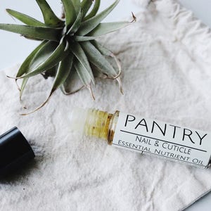 May include: A roll-on bottle of Pantry Nail & Cuticle Essential Nutrient Oil with a black cap, on a white cloth. The label displays the brand name and product details. A small green plant is in the background.