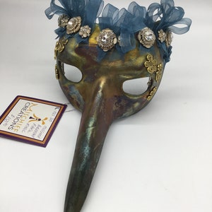 May include: A masquerade mask with a long, curved nose. The mask is painted in shades of brown, green, and gold. It is decorated with blue ribbons, rhinestones, and gold accents. The mask is labeled "Mischlee Creations".