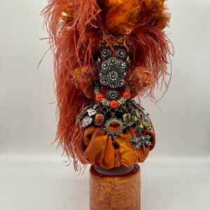 May include: A decorative art piece featuring a fabric head adorned with orange feathers, silver embellishments, and a necklace of orange beads. The head sits atop a cylindrical, textured wooden base, creating a unique and artistic display.
