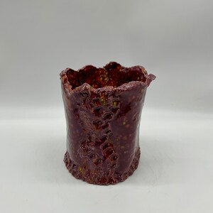 May include: A cylindrical ceramic vessel with a deep burgundy glaze and a glossy finish. The surface features a textured, uneven relief pattern with visible flecks of yellow and orange throughout. The rim and base have a jagged, organic edge.