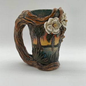 May include: A ceramic mug featuring a sculpted handle resembling a tree branch. The exterior displays a painted landscape of a swamp with trees and water at sunset. Three-dimensional white magnolia flowers are attached to the upper side.