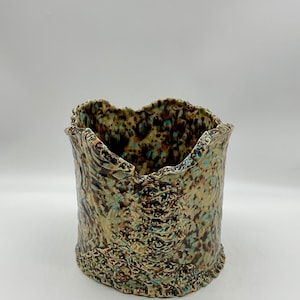 May include: A cylindrical ceramic vessel featuring a highly textured, uneven rim and a mottled glaze finish. The surface displays a complex pattern of brown, tan, and light blue speckles and streaks, creating a rugged, organic appearance throughout.