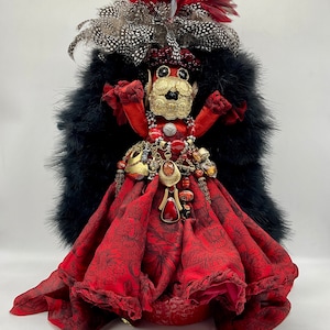 Può includere: Una bambola rossa e nera con un copricapo piumato, un vestito rosso e una collana di perline dorate e rosse. La bambola è in piedi su una base rossa.