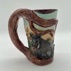Peut inclure: Mug en céramique avec une anse en forme de branche d'arbre. La tasse représente un sanglier dans un cadre naturel, avec un schéma de couleurs turquoise et marron. La poignée est marron foncé.