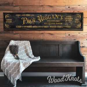 May include: A vintage-style wooden sign with black paint and gold lettering advertising "Prof. Higgan's Traveling Medicine Show". The sign reads "Come one come all, Tonight at Dusk, Cures Ailments in Days, See it Live, Elixirs & Ointments - Snake Oils & Tonics - Miracle Cures & Remedies".