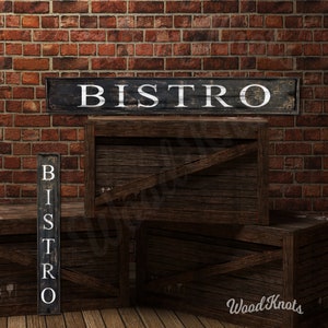 May include: Two wooden signs with the word "BISTRO" in white lettering on a black background. The signs are leaning against wooden crates. The crates are stacked on top of each other. The background is a brick wall.