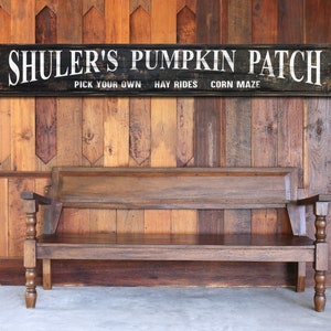 May include: A wooden bench with a dark finish sits in front of a wooden wall. Above the bench is a black sign with white text that reads "SHULER'S PUMPKIN PATCH" and other text below. The sign is weathered.