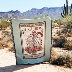 May include: A light blue woven throw blanket with a tarot card design featuring skeletons, flowers, and the words "THE LOVERS." The blanket has a fringed edge and is displayed outdoors with a desert landscape background.