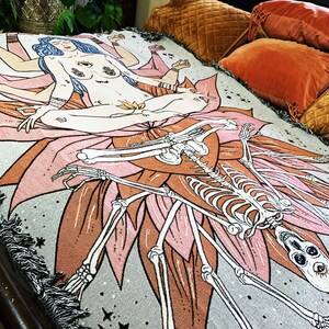 May include: A woven tapestry throw blanket with a gray background featuring a pink lotus flower with a skeleton figure and a woman with multiple arms sitting on the flower. The woman has blue hair and a third eye. The background is a dark gray with white stars.