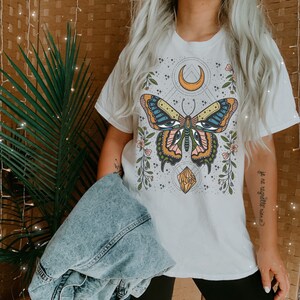 May include: White t-shirt with a colorful butterfly graphic, a crescent moon, and a crystal. The text "je ne regrette rien" is printed on the right side of the shirt.