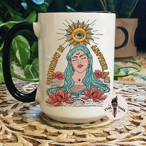 May include: White ceramic mug with a black handle. The mug features a colorful illustration of a woman with long blue hair, a sun with an eye, and the text "Tomorrow is another day".
