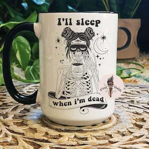 May include: White ceramic mug with a black handle. The mug has a black rim and a black and white illustration of a skeleton with a third eye and wearing a sleep mask. The text "I'll sleep when I'm dead" is printed on the mug.