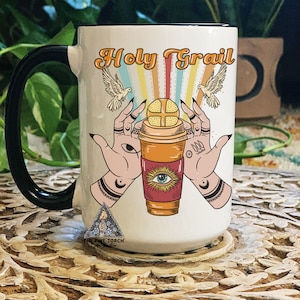 May include: White ceramic coffee mug with a black handle. The mug features a colourful illustration of two hands holding a coffee cup with a red and orange design. The text "Holy Grail" is printed in black above the illustration.