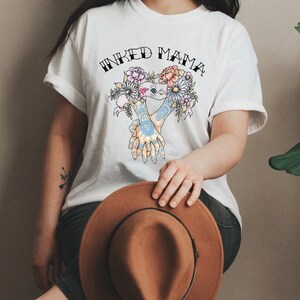 Pu&ograve; includere: T-shirt bianco con la scritta "INKED MAMA" sopra una grafica floreale e di mani colorata. Un cappello marrone &egrave; tenuto in grembo. Il design della maglietta presenta un cuore e fiori.