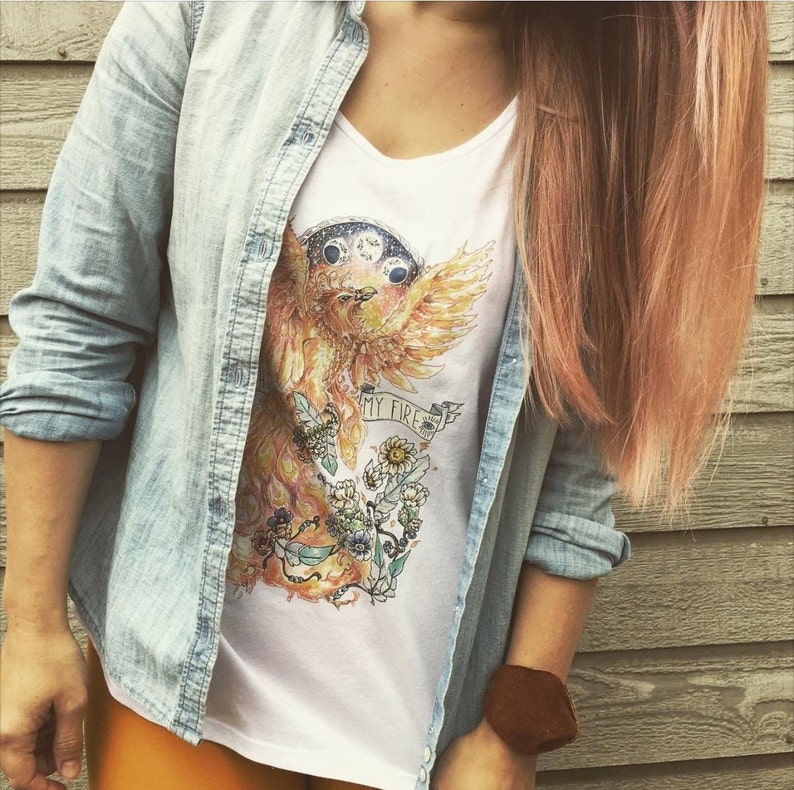 May include: White t-shirt with a watercolor illustration of a phoenix rising from flames. The phoenix has a crown of moons and the text "My Fire" is written on a banner.